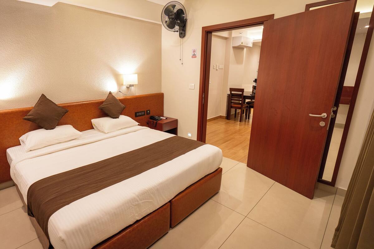 A hotel room with a large bed, bedside tables, a wall-mounted fan, and a door leading to a dining area.