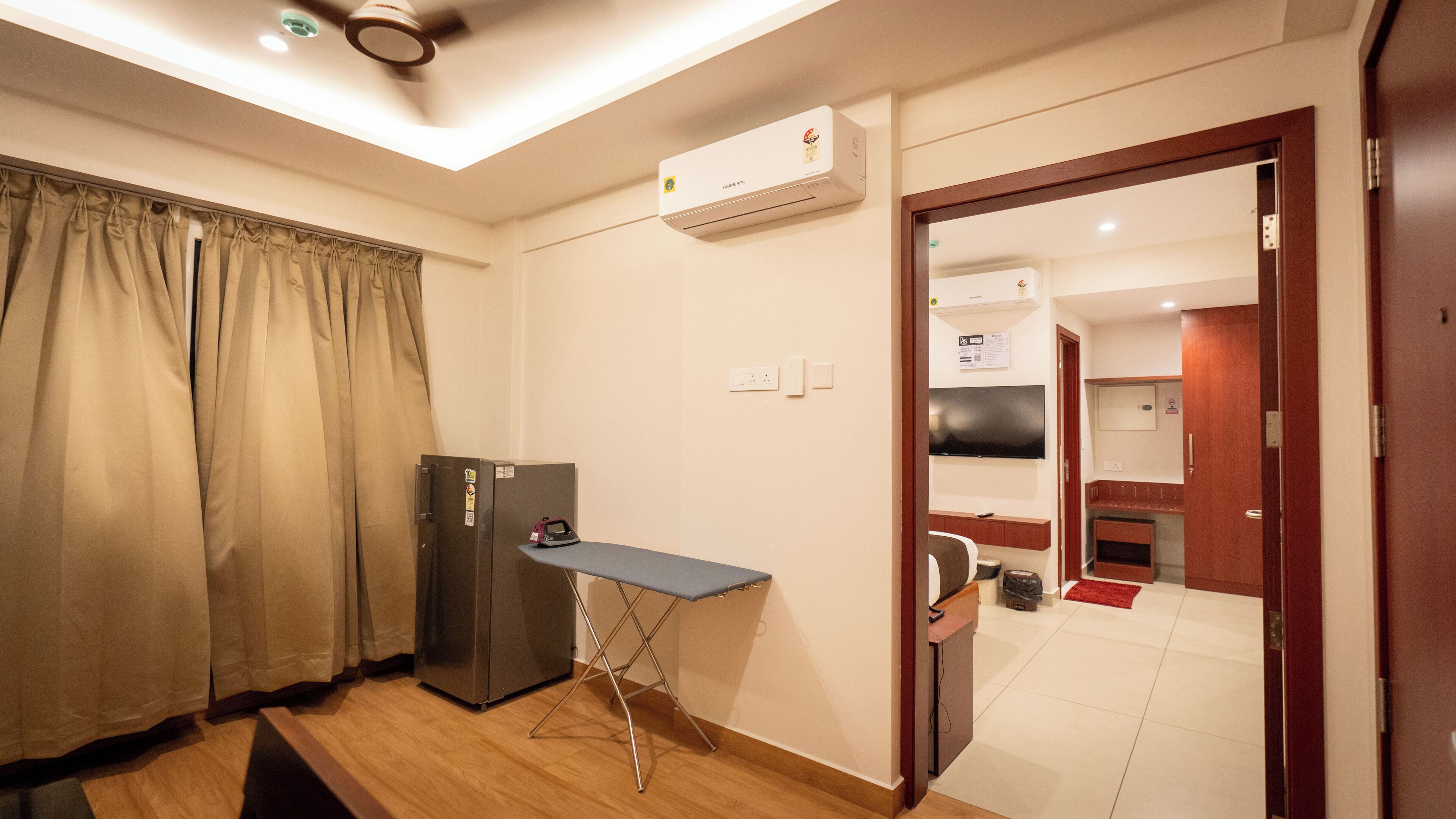 A compact hotel room with a kitchenette, a small ironing board, and a ceiling fan.