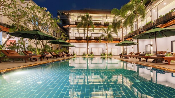 A swimming pool with lounge chairs and umbrellas, surrounded by greenery and a multi-story building with balconies. - The Sears Residence (Siem Reap)