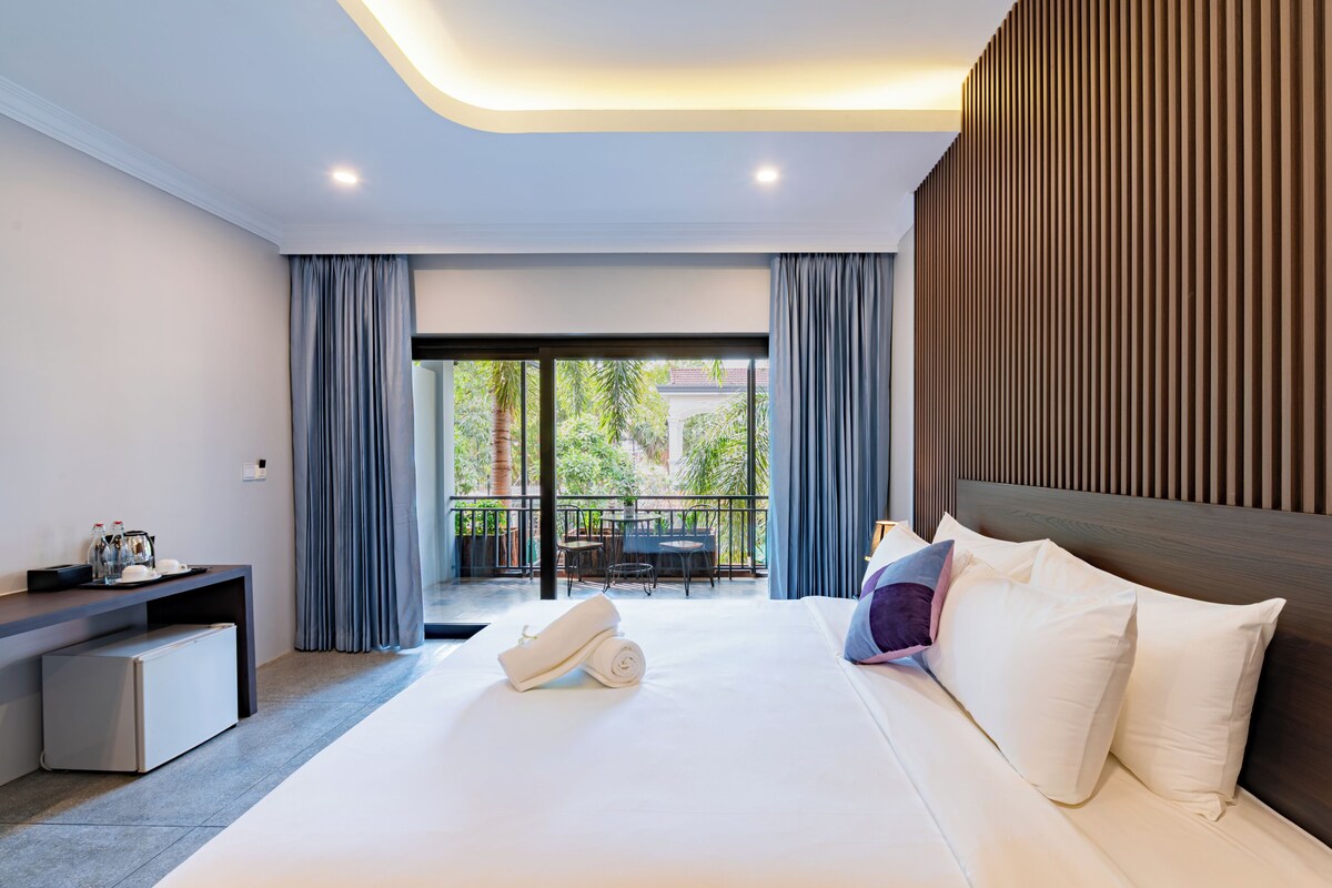 A modern hotel room with a large bed, a desk with a computer, and a balcony with a table and chairs.