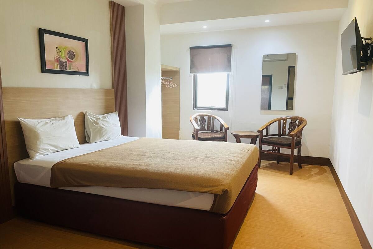 A hotel room with a bed, a chair, a small table, a TV, and a window with a curtain.