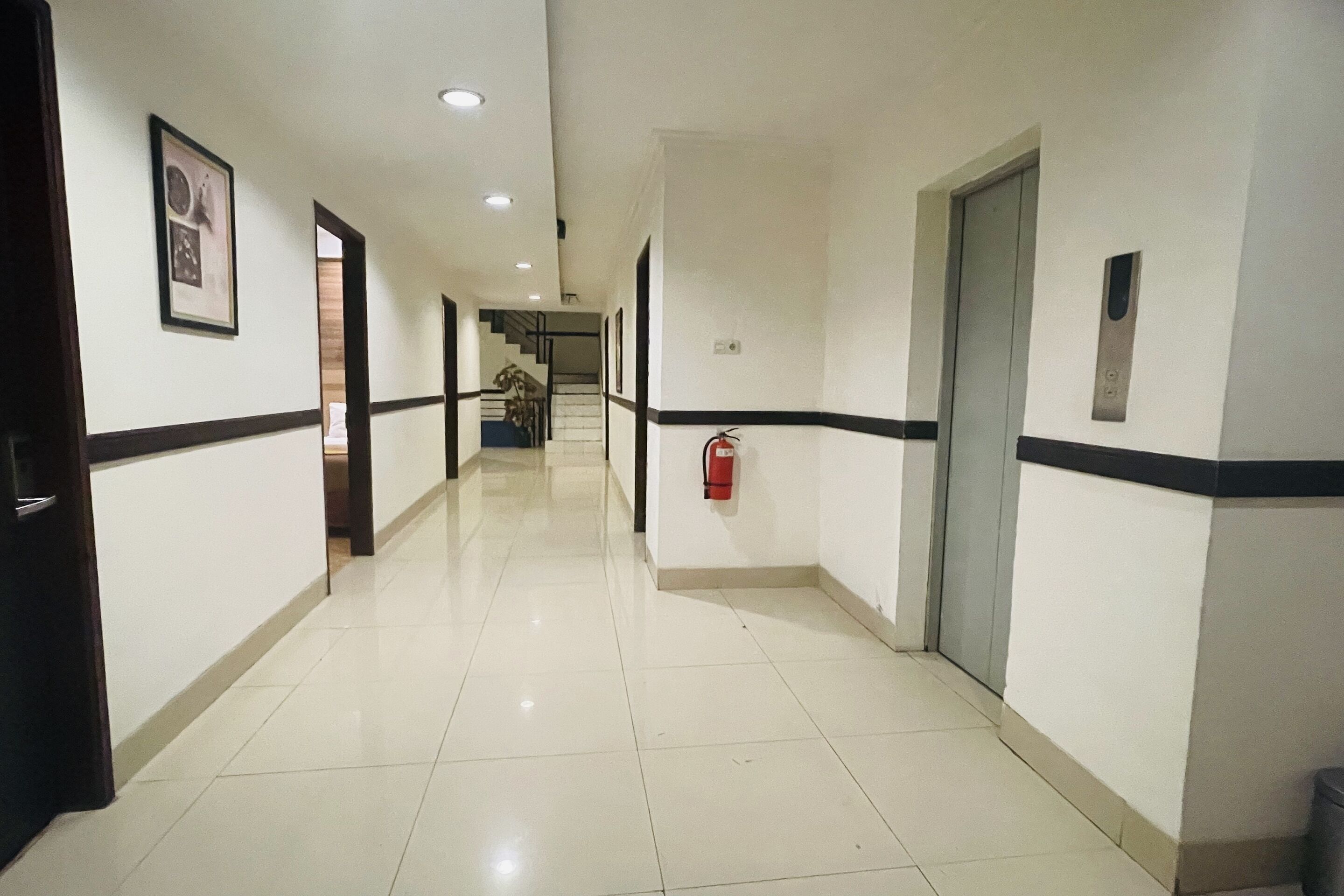 Un pasillo limpio y bien iluminado con paredes blancas, detalles negros y piso de baldosas. Hay puertas a ambos lados, un extintor de incendios montado en la pared y una escalera al fondo.