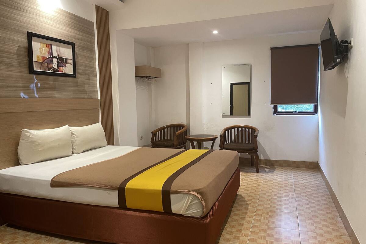 A hotel room with a bed, a small table, two chairs, a TV, and a window with a blind.
