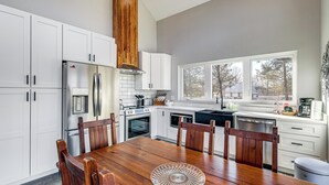 Una cocina moderna con armarios blancos, electrodomésticos de acero inoxidable, una mesa de comedor de madera y un ventanal grande.