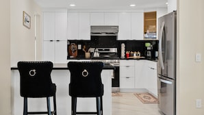 A modern kitchen with white cabinets, a stainless steel refrigerator, two black bar stools, and a black countertop. - Relax & Unwind: Pool, Jacuzzi & Private Backyard (Rowland Heights)