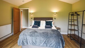 Quarto com teto inclinado, cama com manta cinza, duas luzes fixadas na parede, porta de madeira e estante de metal.