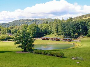 Un paysage rural avec un étang, des champs verdoyants et une rangée de cabanes en bois.