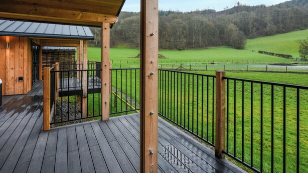 Um deck de madeira com guarda-corpo de metal, área coberta e vista para um campo verde e árvores.