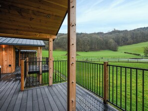 Een houten terras met een metalen hekwerk, een overdekt gedeelte, en uitzicht op een groen veld en bomen.