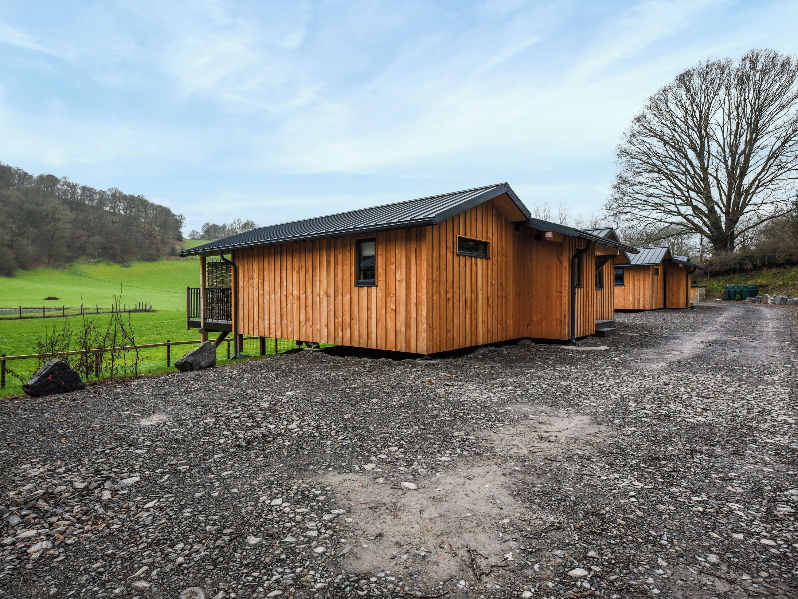 A wooden building with a sloped roof, a gravel driveway, and a fenced green field in the background.