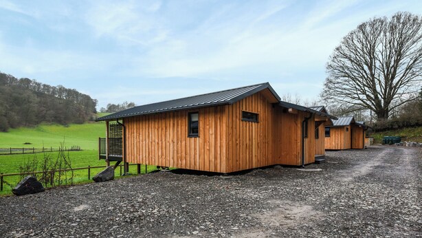 A wooden building with a sloped roof, a gravel driveway, and a fenced green field in the background. - Lakeside Lodge - UK50627 (Welshpool)