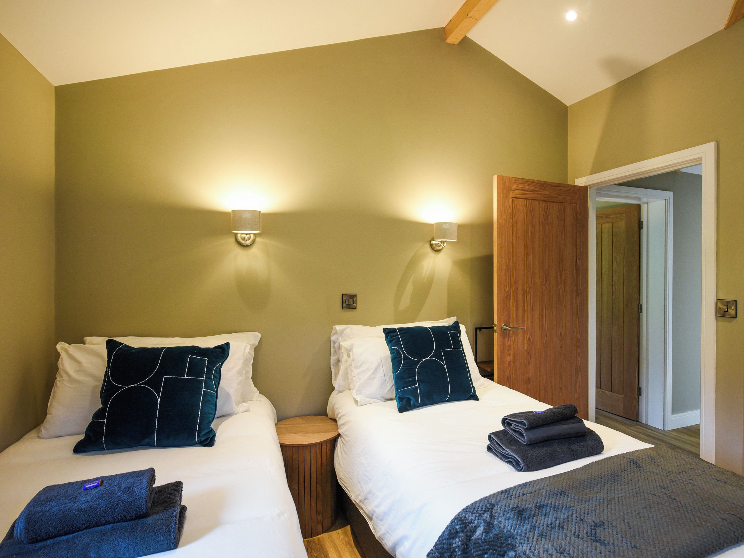 Ein Hotelzimmer mit zwei Betten, jeweils mit Kopfkissen und Decke, einem runden Nachttisch aus Holz und einer Tür zu einem weiteren Raum.