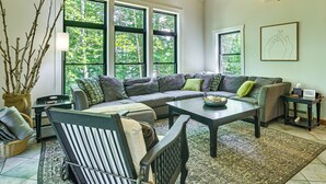 Una sala de estar con sofá, sillón, mesa de centro y un gran ventanal con vista a árboles.
