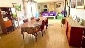 Spisestue med et rundt bord og stole, et fjernsyn i baggrunden og en sofa i stuen.