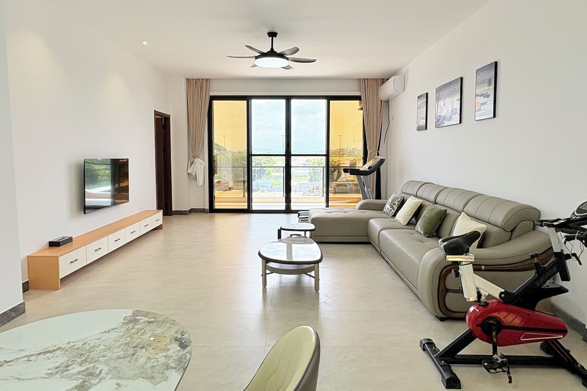 A modern living room with a sofa, TV, exercise bike, and a balcony with a view.