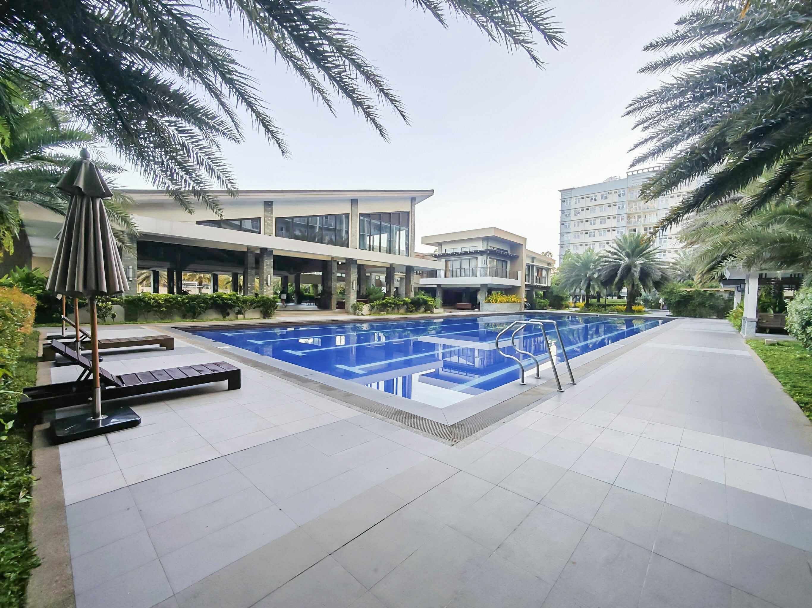 A swimming pool with lounge chairs and a covered seating area, surrounded by palm trees and modern buildings.