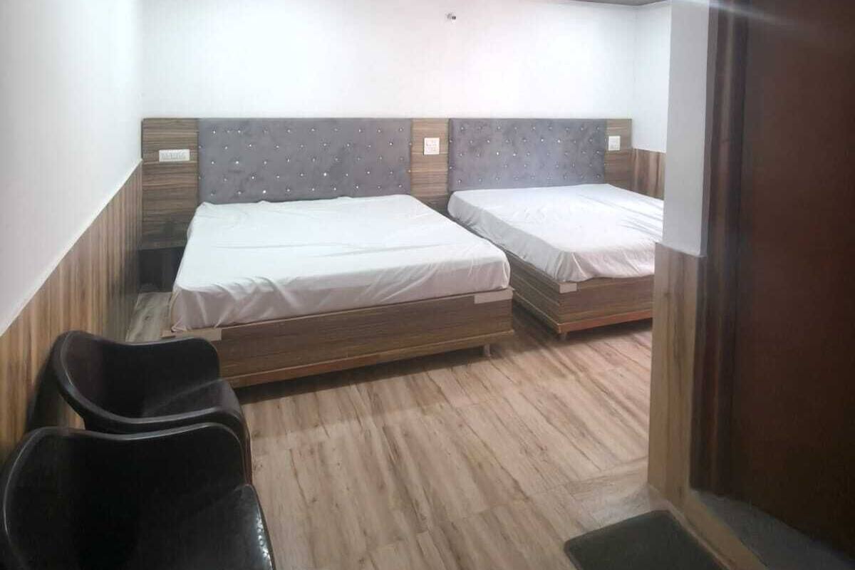 A room with two beds, wooden flooring, a ceiling with recessed lighting, and a door.
