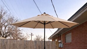 Een ronde glazen tafel met een zwart metalen onderstel, omringd door vier witte plastic stoelen met zwarte voetensteunen. Een parasol zorgt voor schaduw boven de tafel.