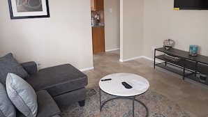 Een woonkamer met een grijze bank, een ronde salontafel en een televisie aan de muur.