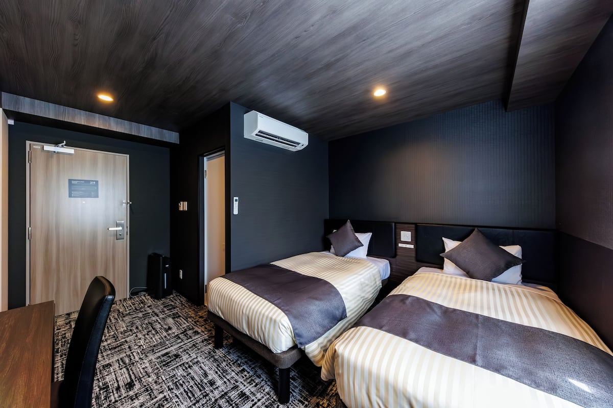 Habitación de hotel con dos camas, un escritorio, una silla, un equipo de aire acondicionado y una puerta con un cartel.