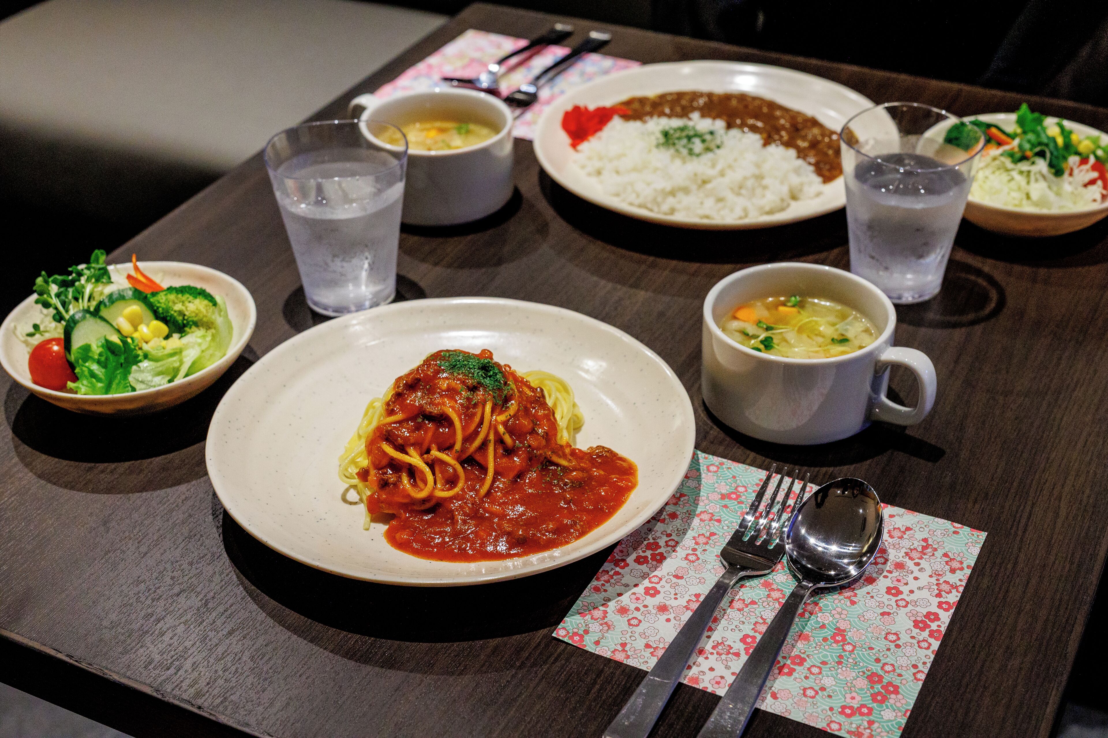 A table with various dishes, including pasta, rice, and vegetables, along with cups of water and a spoon.