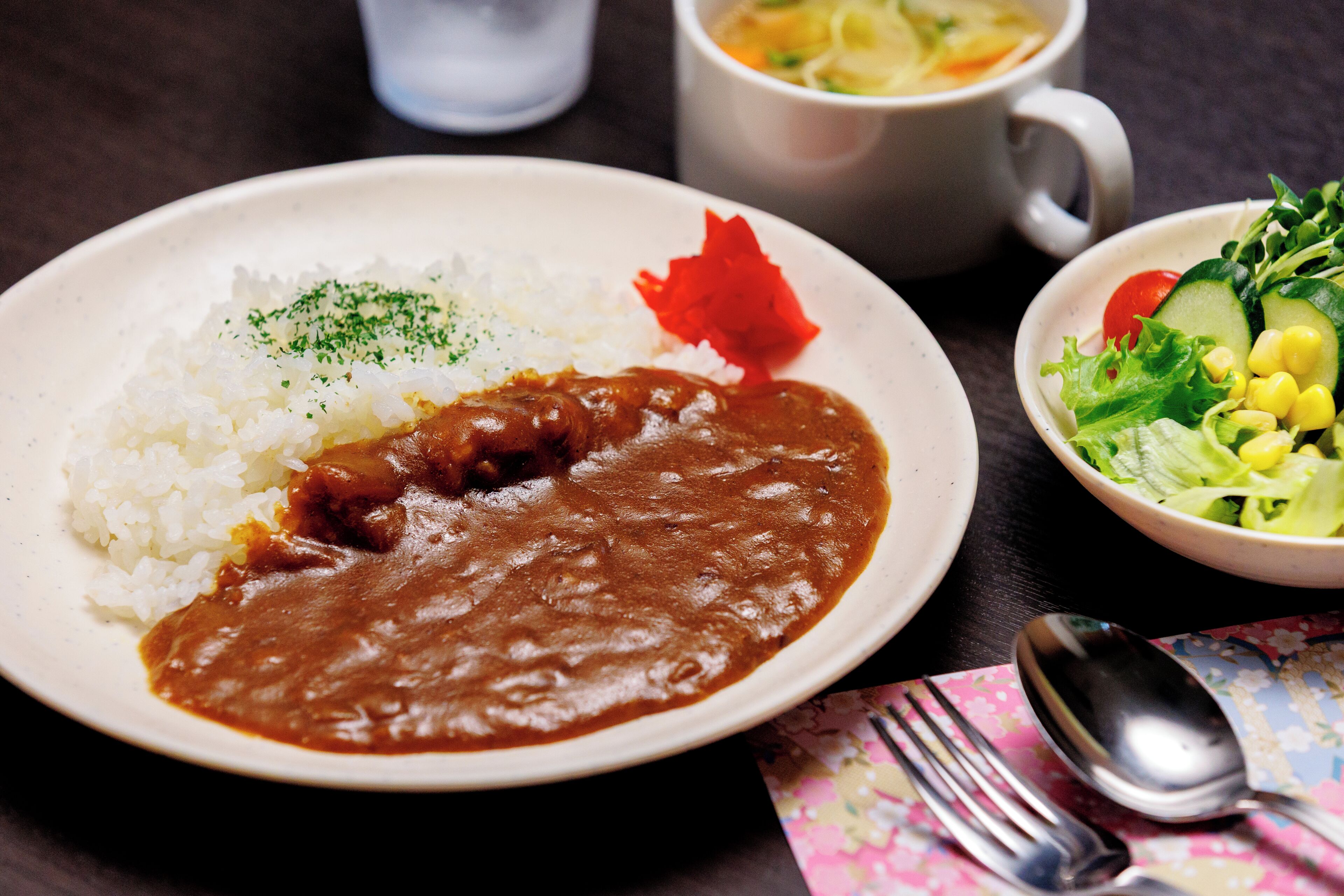 A plate of white rice with brown sauce, a bowl of salad, and a cup of soup on a table.