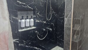 Et badeværelse med væg i sort marmor, en bruser med håndbruser og en hylde med toiletartikler.