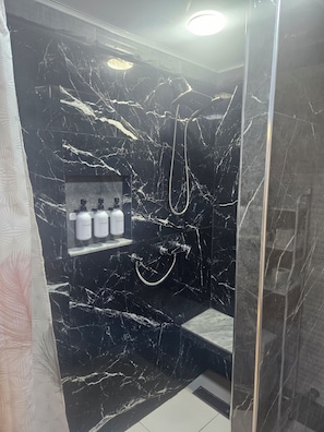 Un bagno con parete in marmo nero, doccia con soffione a mano e ripiano con articoli da toilette.