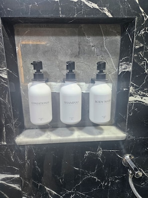Uno scaffale con cornice in marmo e tre bottiglie bianche etichettate con dopobarba, shampoo e bagnoschiuma.