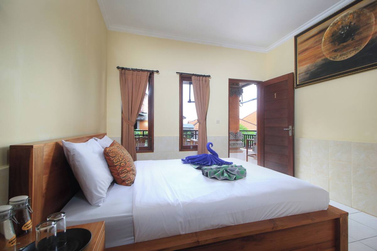 Una cama bien hecha con sábanas blancas, cabecera de madera y un cojín decorativo. Hay dos ventanas con cortinas, y un cuadro de una escena celestial cuelga en la pared.
