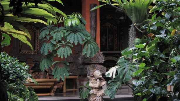 A traditional garden with stone statues, large leafy plants, and a wooden bench. - Tusantha Ubud A Trishula Collection (Ubud)