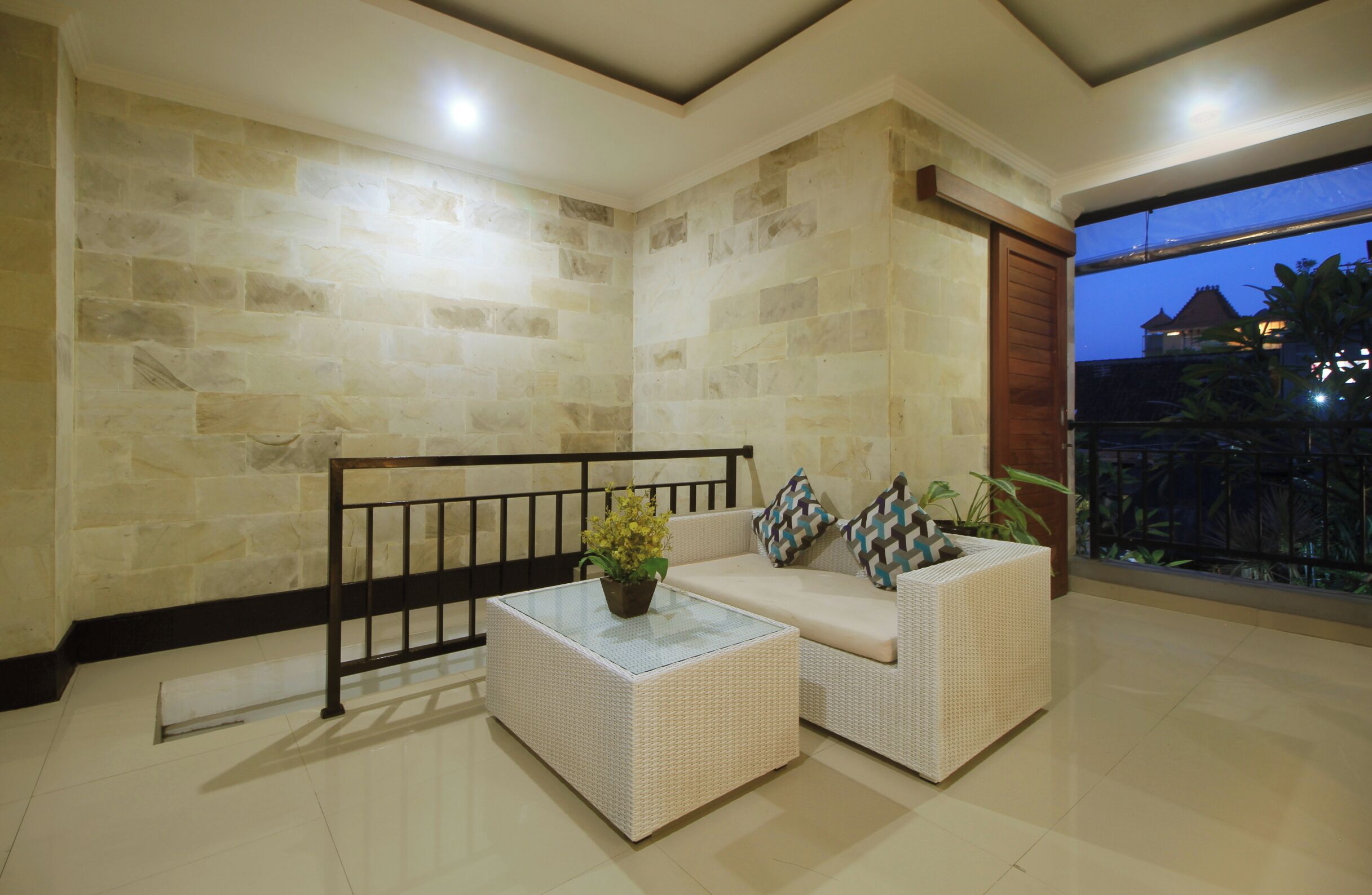 Quarto de canto com paredes de pedra, mesa de centro de vidro, sofás de vime e vista para um edifício ao entardecer.