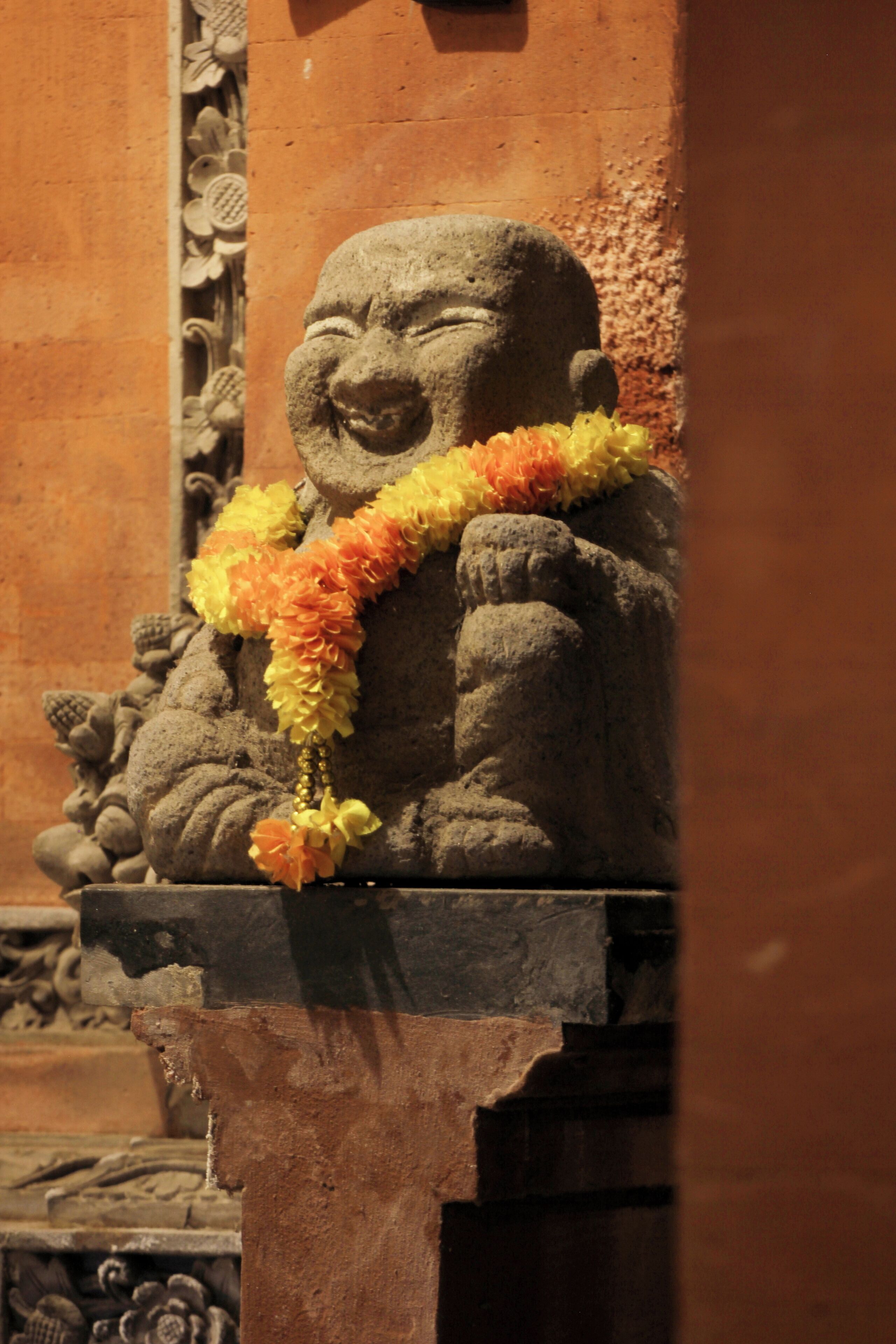 A stone statue of a laughing Buddha adorned with a garland, set against a textured wall.
