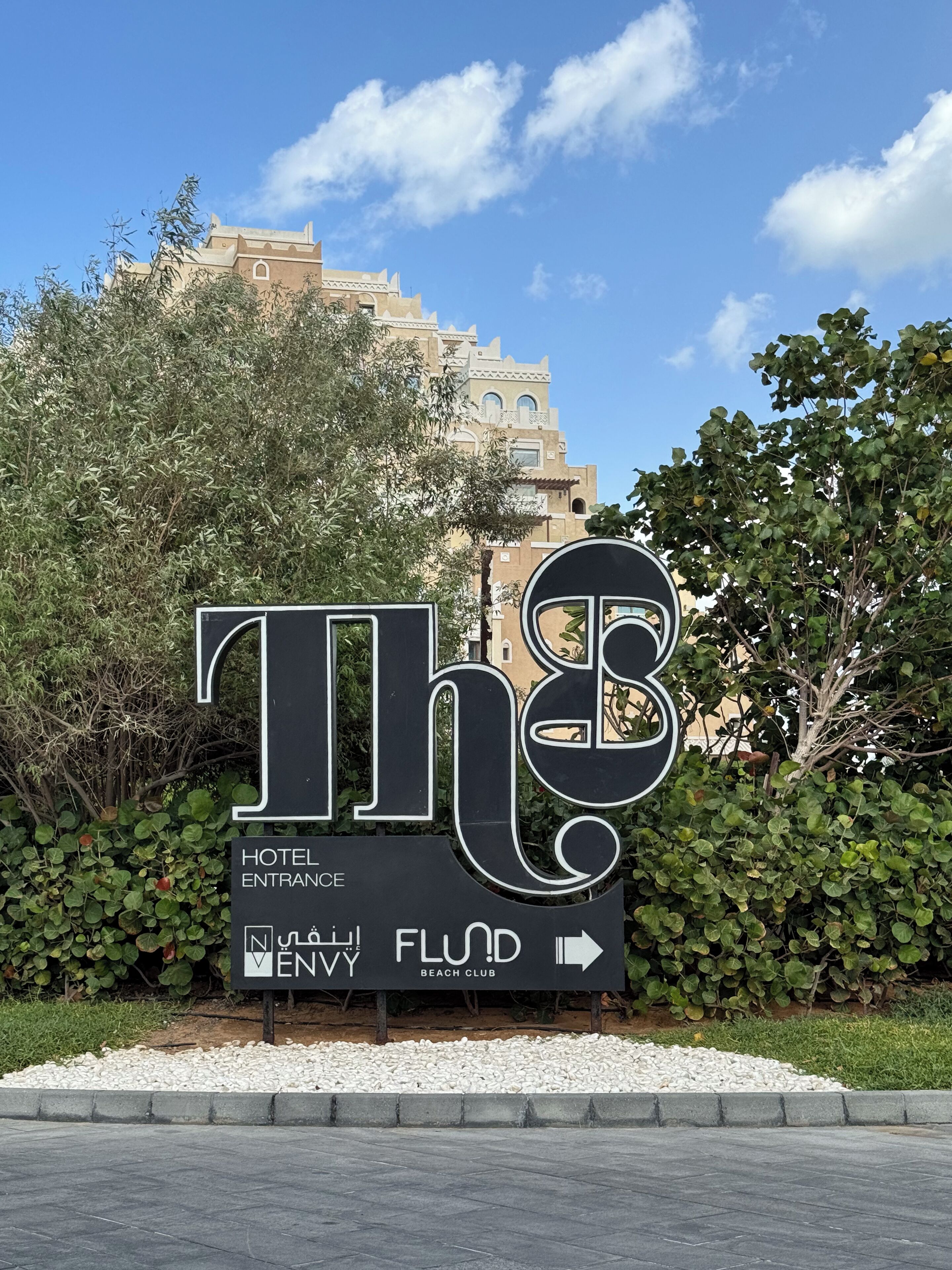A hotel entrance sign with "The" in stylized letters, "HOTEL ENTRANCE" and "ENVY FLOR BEACH CLUB" text, and an arrow pointing right.