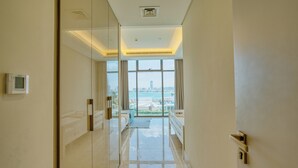 A modern hotel corridor with a glass partition, a bed, a small seating area, and a view of the cityscape and water. - PVH - The8 - Opulent 3BR - Beach Access (Dubai)