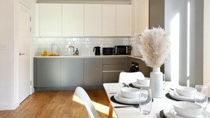 A modern kitchen with white cabinetry, a dining area with a white table set for four, and a decorative vase with pampas grass. - The Central Mount-Balcony-Gym-Parking (Manchester)