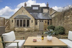 Une maison en pierre dotée d’une baie vitrée, d’une porte et d’un petit jardin avec une table, des chaises et une bouteille de vin.