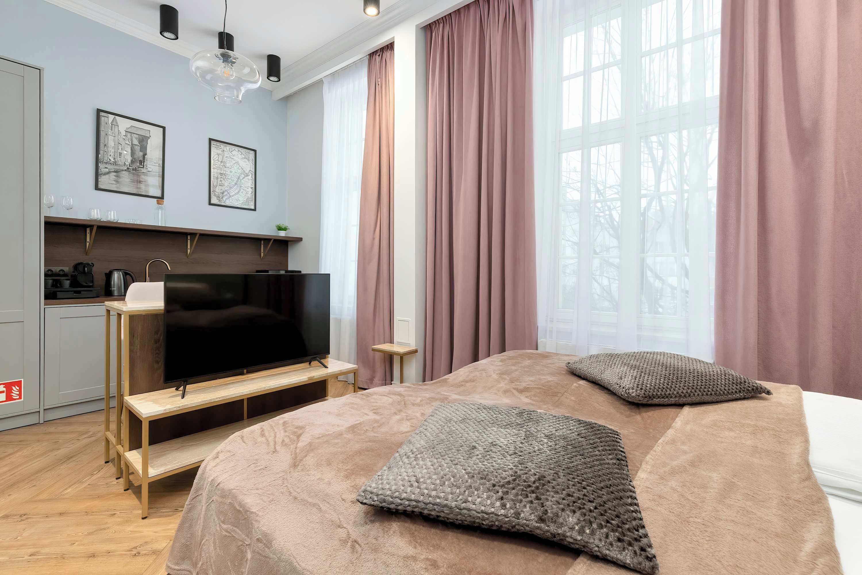 Een moderne hotelkamer met een groot bed, een flatscreen televisie, een kleine kitchenette en grote ramen met gordijnen.