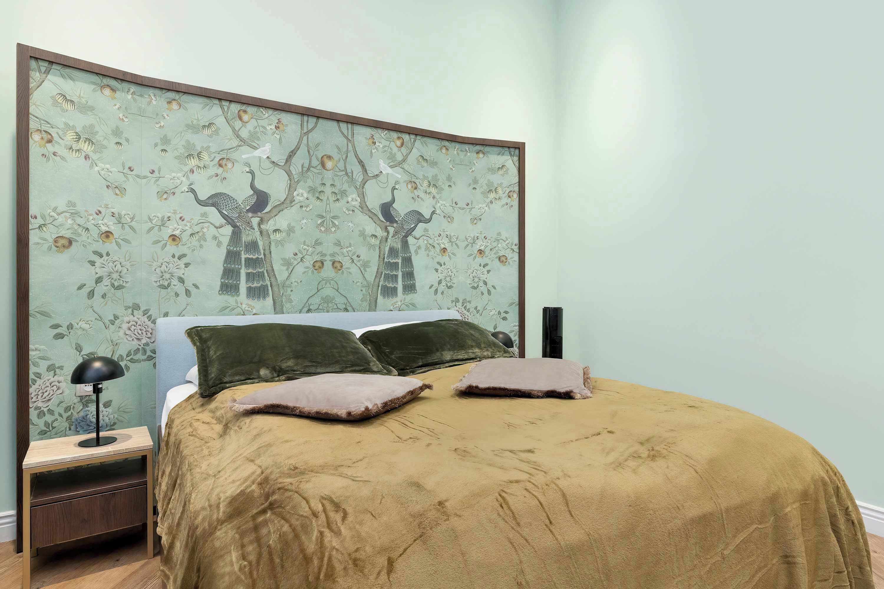 A bedroom with a bed, pillows, a nightstand, a lamp, and a wall with a floral and tree mural.