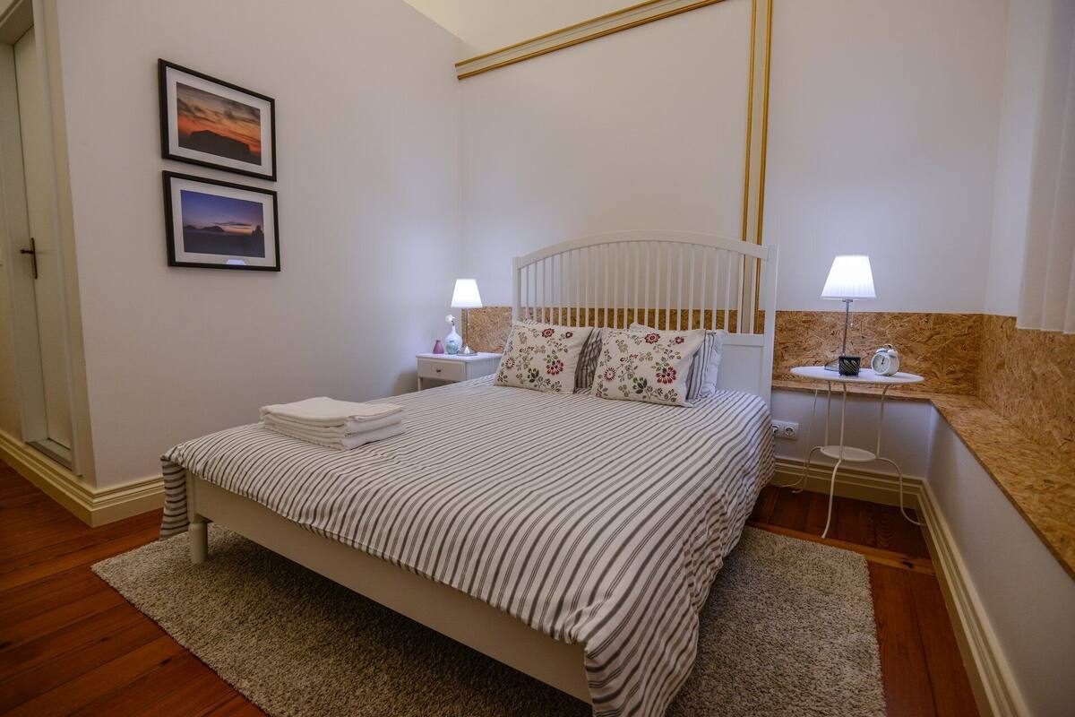 A neatly arranged bedroom with a bed, two bedside tables, a nightstand, a wall-mounted lamp, and two framed pictures.
