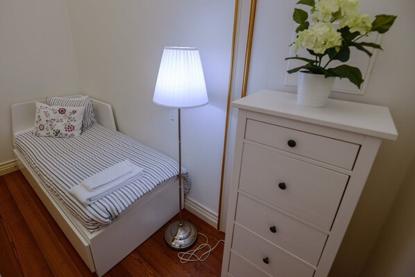 Una cama bien hecha con ropa de cama a rayas y un cojín floral, una lámpara de noche, un tocador blanco con cajones y una planta en maceta encima.