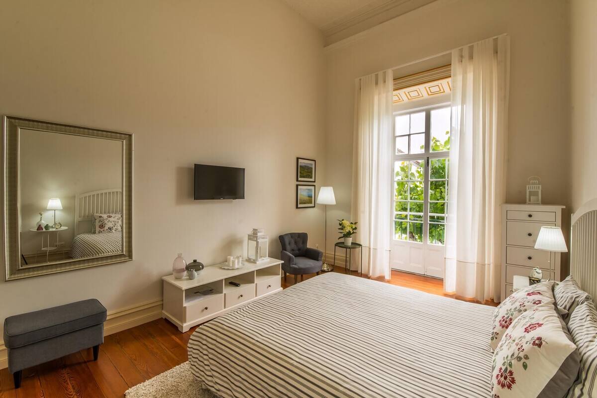 A bedroom with a large bed, a TV, a chair, a desk, and a window with curtains.