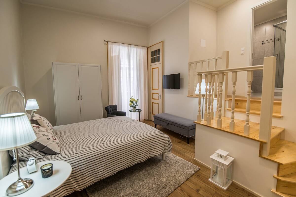 A bedroom with a bed, bedside lamps, a desk, a chair, a TV, a staircase, and a wardrobe.