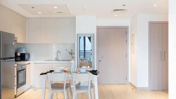 A modern kitchen with white cabinetry, a stainless steel refrigerator, a coffee maker, and a dining area with two white chairs and a table. - Bloomfields Iconic 1BR Waters Edge (Abu Dhabi)