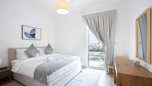 A modern bedroom with a large bed, a bedside table, a desk, a window with curtains, and a painting on the wall. - Bloomfields Iconic 1BR Waters Edge (Abu Dhabi)