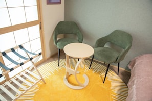 Quarto com tapete amarelo estampado, duas cadeiras verdes, uma mesa redonda pequena e um berço com estrutura de madeira e tecido azul.
