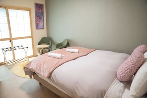 Uma cama bem arrumada com manta e travesseiros cor-de-rosa, uma mesa redonda pequena, uma cadeira verde e uma janela com vista.