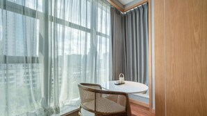 Una habitación moderna con una mesa redonda y dos sillas de mimbre, ventanas amplias con cortinas translúcidas y vistas a edificios.