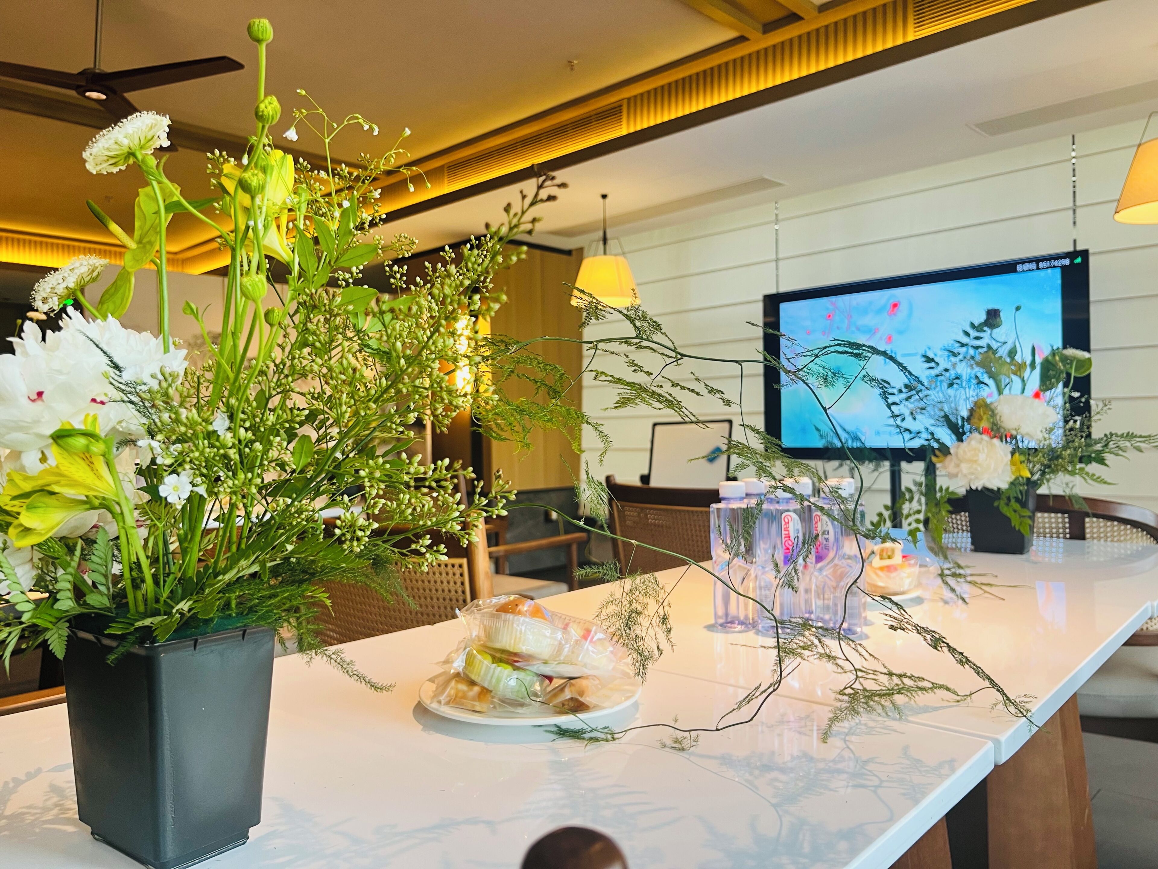 Een hotel receptie met een marmeren balie, een potplant met bloemen, een televisiescherm en een waterdispenser.