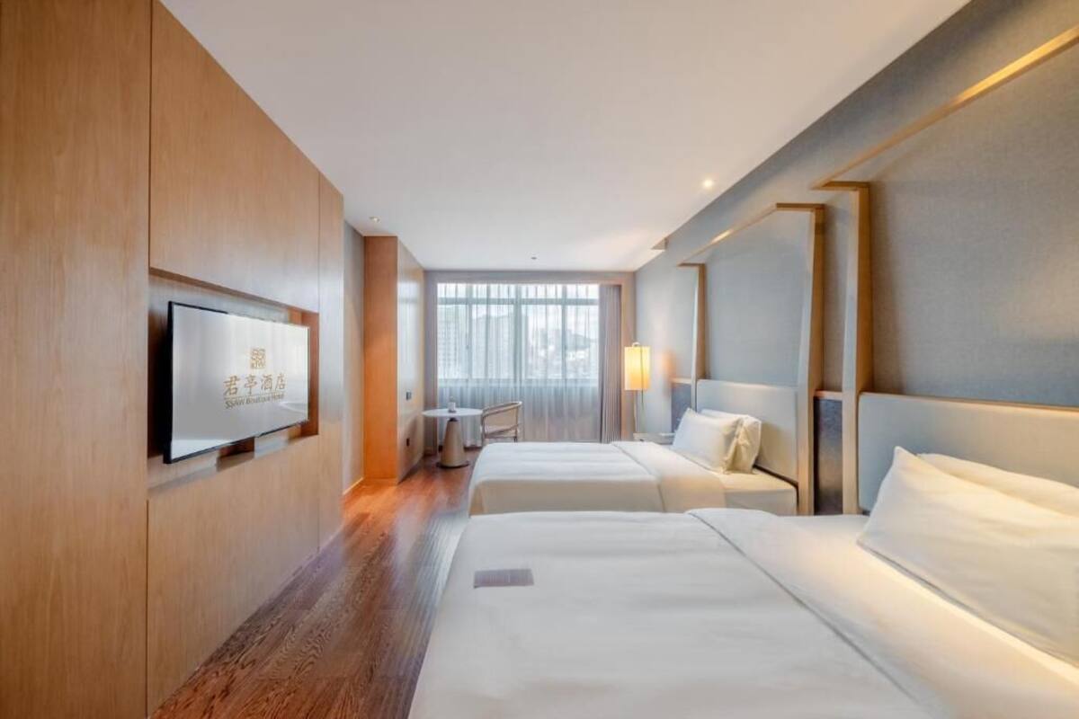 Habitación de hotel con dos camas, un televisor montado en la pared, una mesita y una ventana con cortinas.